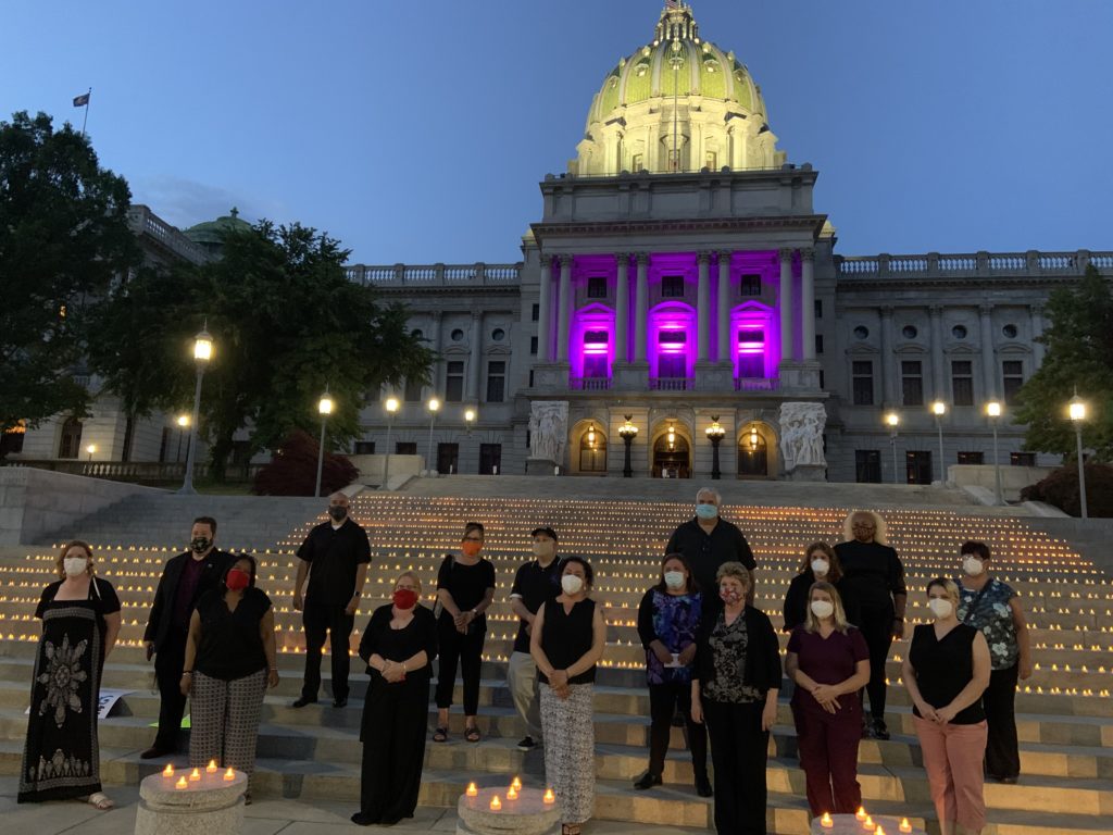 image: union. members dressed in black wearing masks standing in front of thousands of candles laid out on the steps of the Pennsylvania Capitol